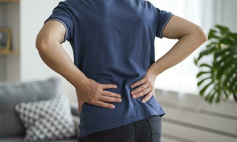 Man stands with back to camera with hands placed on his lower back, slightly bent over in pain.