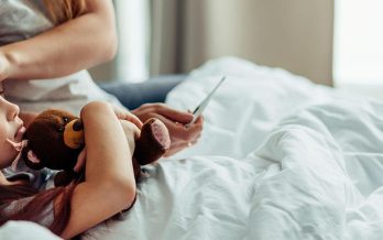 A young child lies in bed holding a teddy bear, with a caregiver checking her forehead and holding a thermometer.