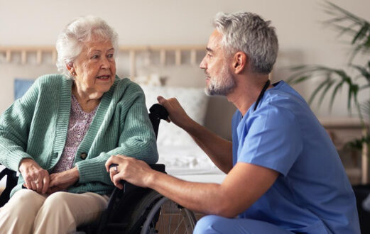 A healthcare worker kneels beside an older woman in a wheelchair, speaking with her warmly.