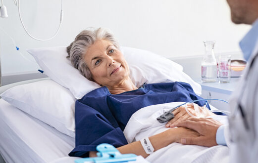 An older patient smiles up at her doctor from her hospital bed.