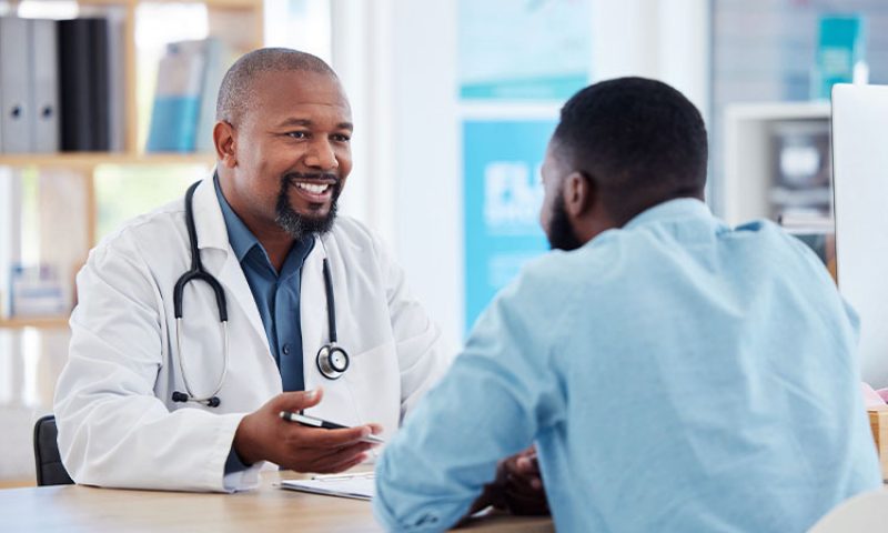 A doctor smiles and talks with a male patient across the desk.