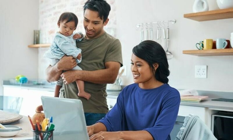 Smiling couple with a young child, the father holding the toddler while the mother types on a laptop in the kitchen.