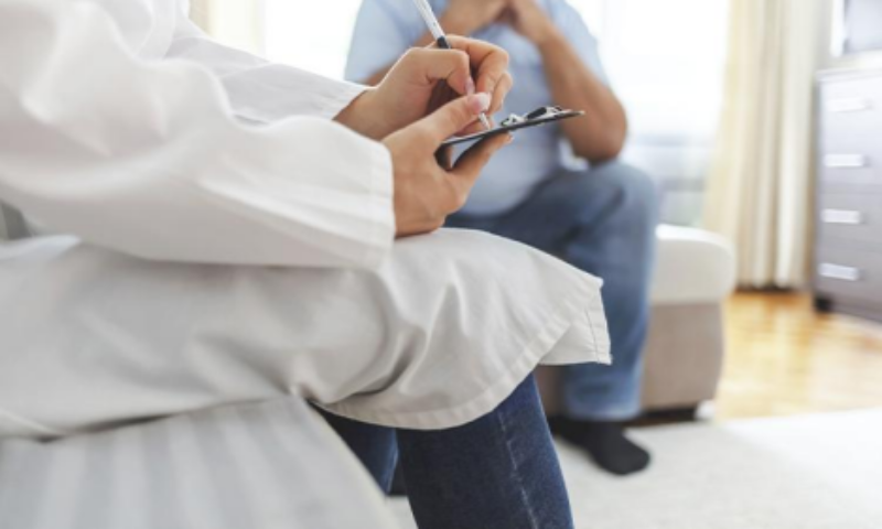 Close-up of a person in a white coat writing on a clipboard while another person sits across from them.