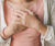 A woman crosses her hands over her chest. She wears a pink tee shirt and a cream cardigan.