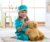 A young girl sits on a rug in her bedroom playing dress up as a doctor. She wears blue scrubs and holds a teddy bear.