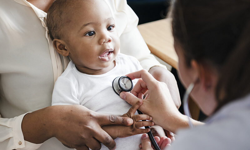 Close up of a smiling baby as a doctor listens to their heart with a stethoscope