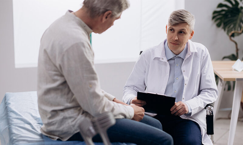 doctor talking to man sitting on hospital chair next to his crutches