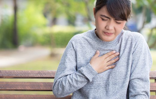 Woman in gray sweatshirt hold hand over upper chest near her throat in discomfort