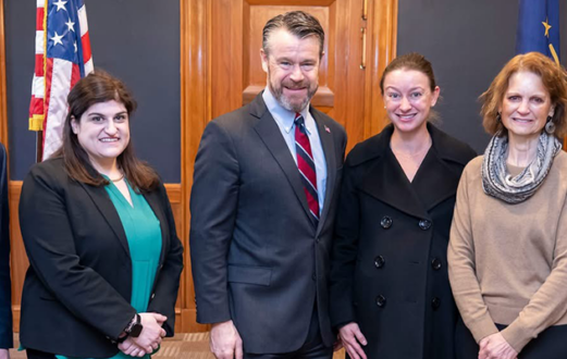 Six people smiling towards the camera and standing together in a board room in front of the American flag
