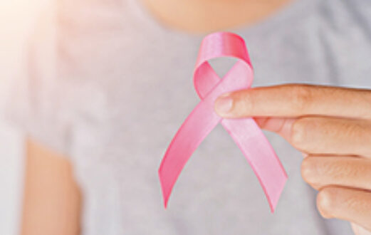 hand holds pink ribbon close to camera, with woman in gray shirt in background