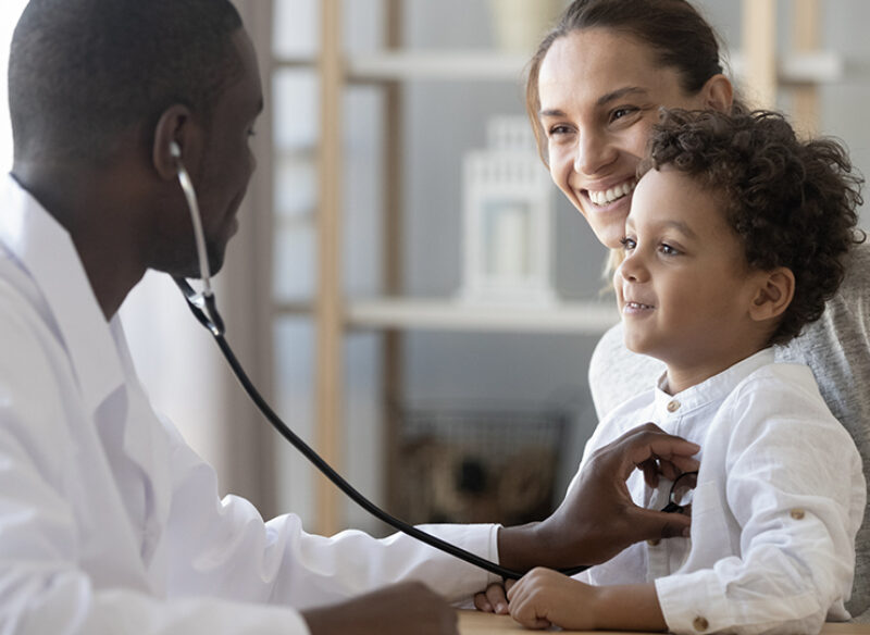 A doctor checks the heart of a young patient sitting on his mom's lap.